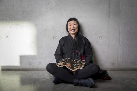 An artist wearing black sits cross-legged on a concrete floor against a concrete wall.