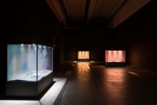 An installation view of a darkened gallery space, in which three glass tanks are filled with brightly lit coloured dust.