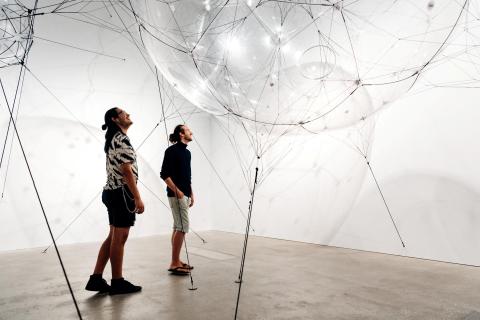 A view of a large bubble-like inflated structure installed in a bright gallery space; two visitors stand underneath looking up into it.
