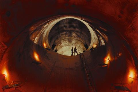 A photograph taken from the bottom of a tunnel underground, with a red glow illuminating the mouth of the tunnel, where small figures stand.