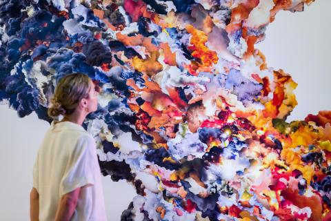 An installation view of a colourful work composed of many plumes of smoke collaged together, with a gallery visitor looking on at left.