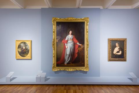An installation view of a very large oil painting in a gold-gilded frame, with a smaller work displayed on either side, on a light blue gallery wall.