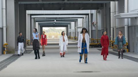 A photograph of seven young people in androgynous contemporary fashions walking through a well-lit industrial space.