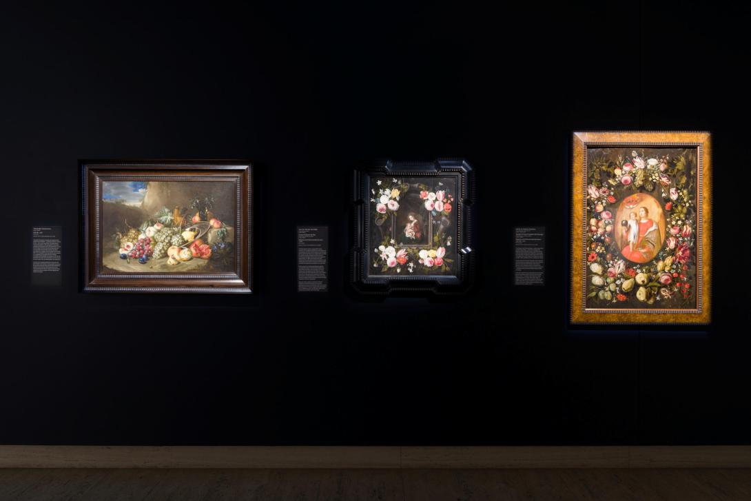 An installation view of a dark gallery space, where the walls are painted black; three still life oil paintings are hung on the wall