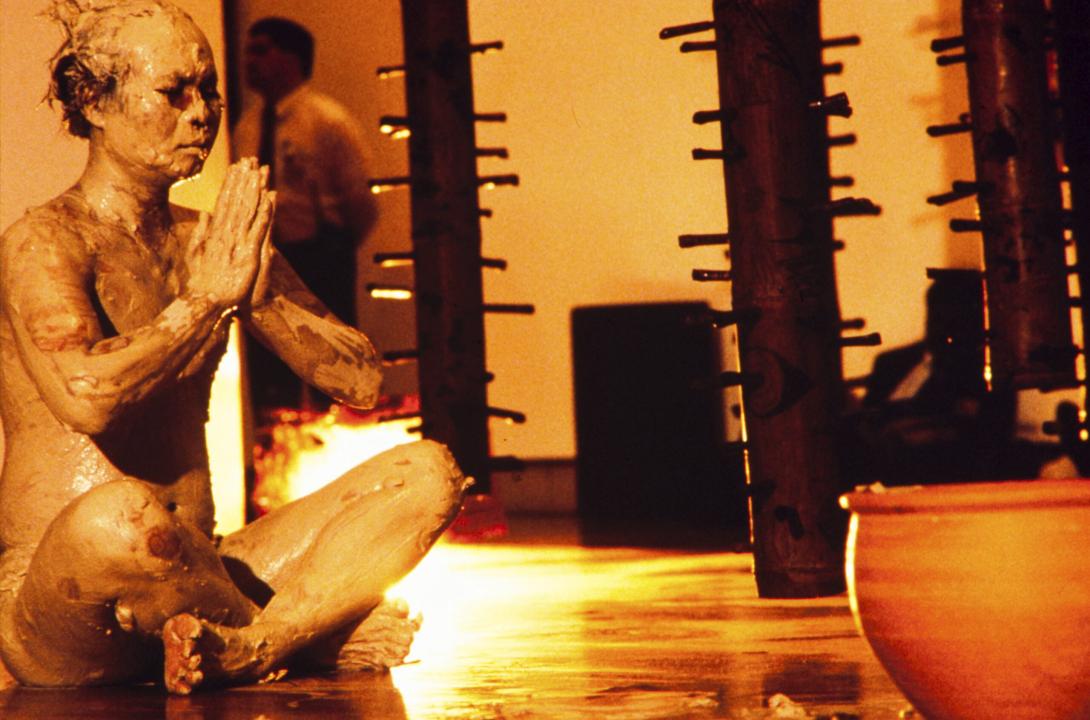 A photograph of a performance artist appearing to meditate, covered in mud or clay, next to an installation of tree-like sculptures.