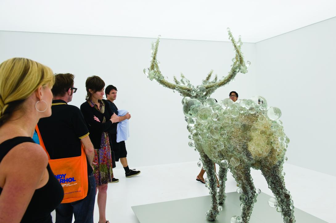 An installation view of a sculpture of a deer that appears to be made from glass bubbles; we see the deer from behind, with onlookers at left.