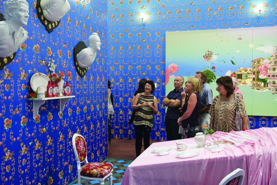 An installation view of an immersive work featuring graphic, electric blue wallpaper with busts hung on the wall, and a long table dressed with a pink tablecloth and propped up at one end.